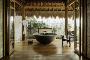 privater aussenbereich überdachte terrasse mit stroh dach an der bambus säule hängt ein bademantel und das alles ist im innenhof auf der terrasse steht eine grosse stein schale als badewanne mit einem wasser einlass aus metall sehr edel im exotischen und 
